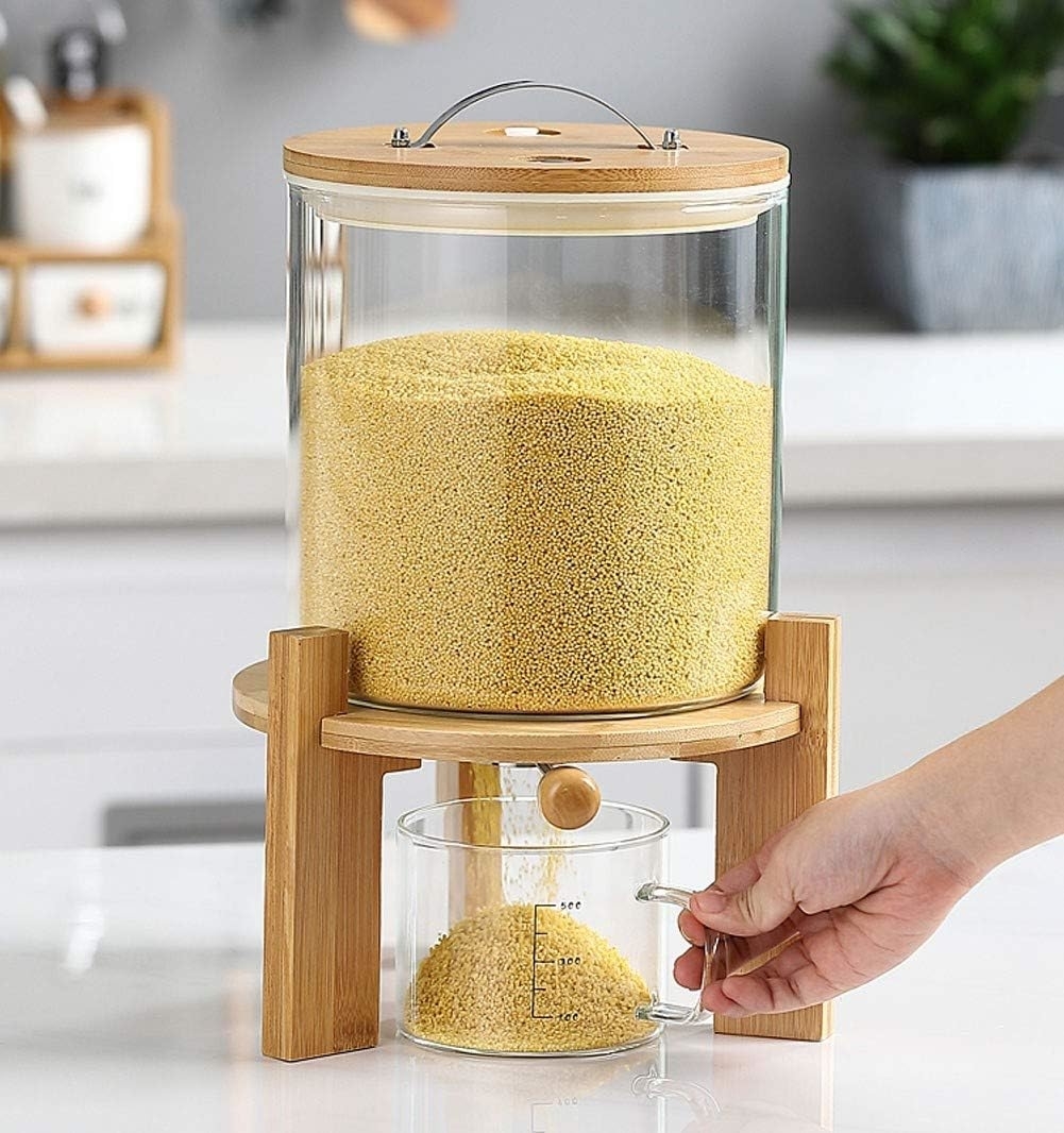 A hand dispenses grains from a large glass and bamboo container into a measuring cup in a modern kitchen