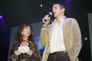 Kathy Griffin and Ashton Kutcher on stage. Kathy holds note cards and wears a velvet jacket. Ashton holds a microphone, dressed in a blazer and light shirt