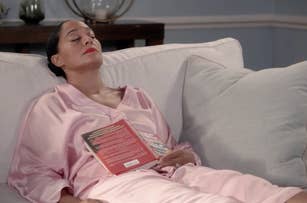 Tracee Ellis Ross relaxes on a couch wearing satin pajamas, with a book resting on her lap, eyes closed