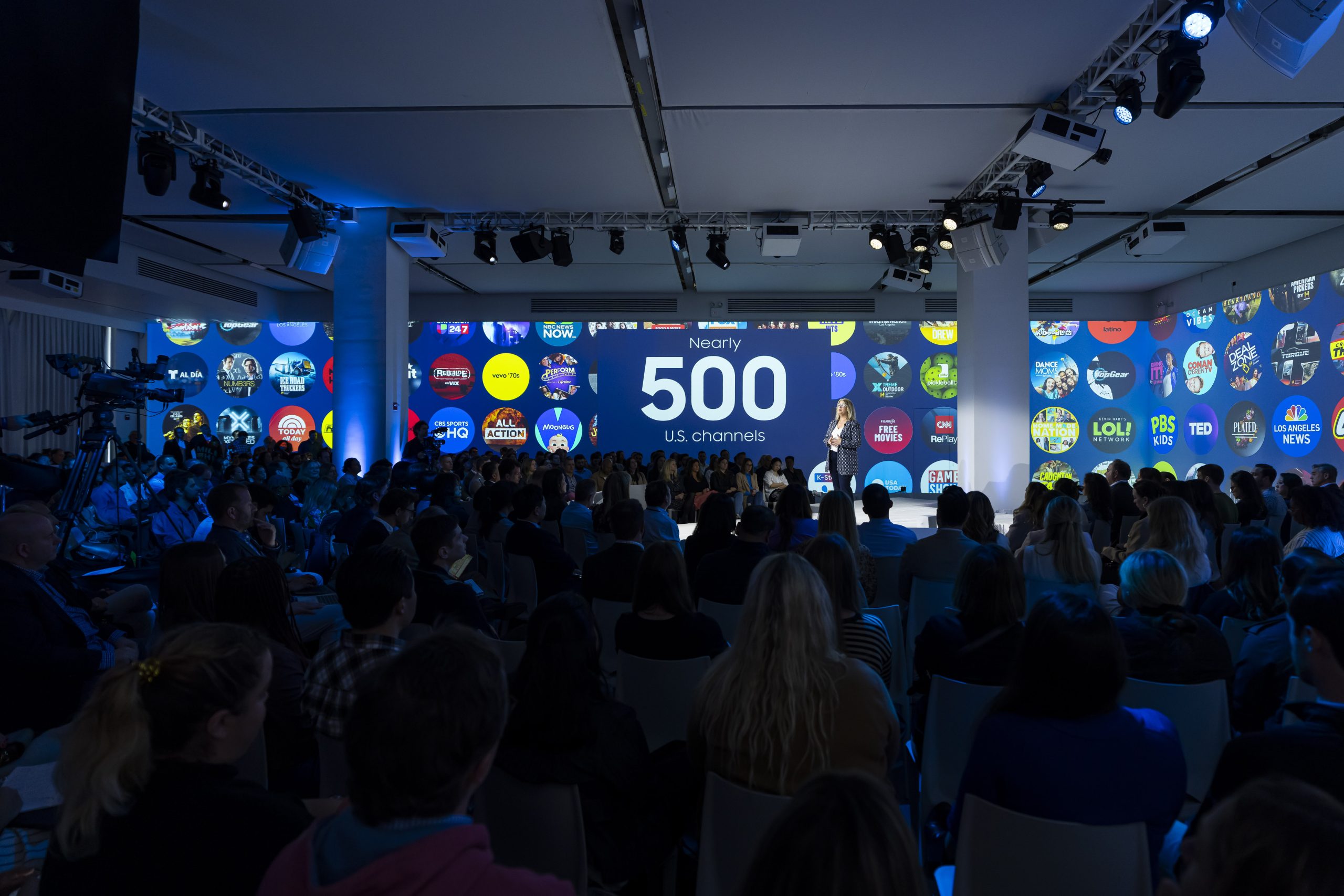 Nearly 500 US channels, logos of TV channels displayed on multiple screens with speaker on stage