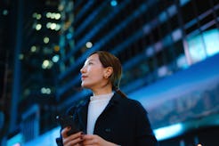 A woman holding a smartphone gazes thoughtfully amidst vibrant city lights at night.