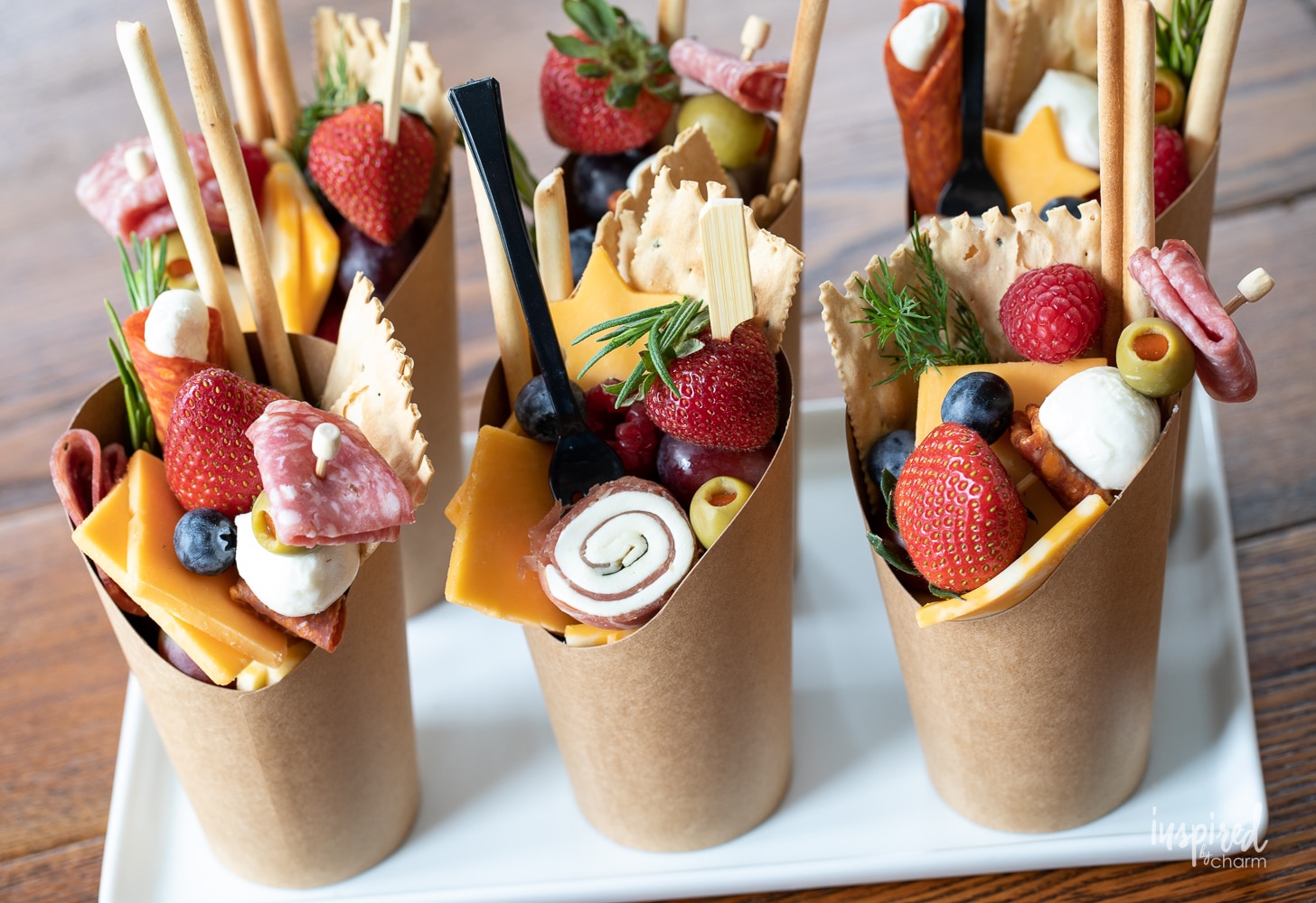 six charcuterie cups filled with meat, cheese, and fruit on a white platter.