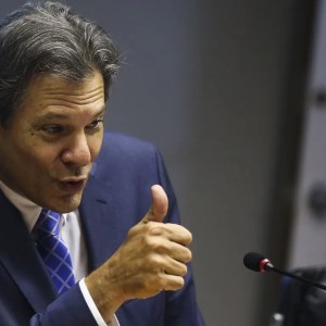 Foto do ministro da Fazenda, Fernando Haddad; ele é um homem branco, de cabelos grisalhos, veste um terno azul marinho e gravata listrada, e faz um sinal de positivo com a mão