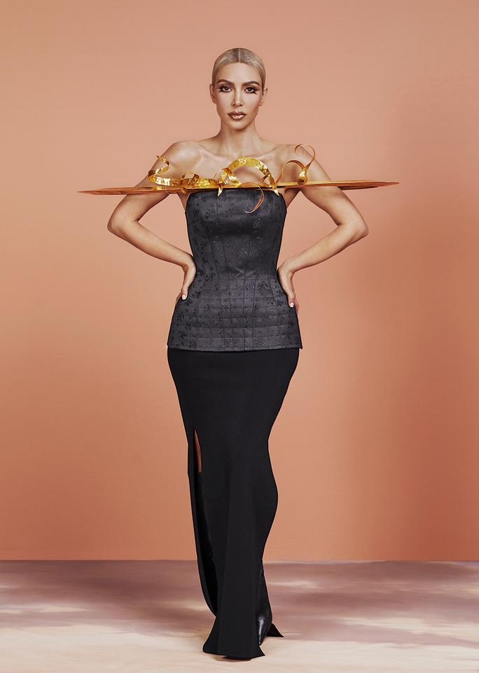 Full-length portrait of Kim Kardashian in front of a dusty salmon seamless background. She wears a floor length black column dress with a brass Saturn-like ring around the top. 