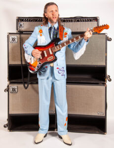 Jimmy Touzel in a blue suit holding an electric guitar in front of multiple amps