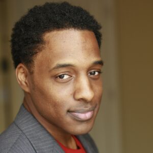 person photographed for headshot in grey tuxedo with red shirt underneath looking to the camera