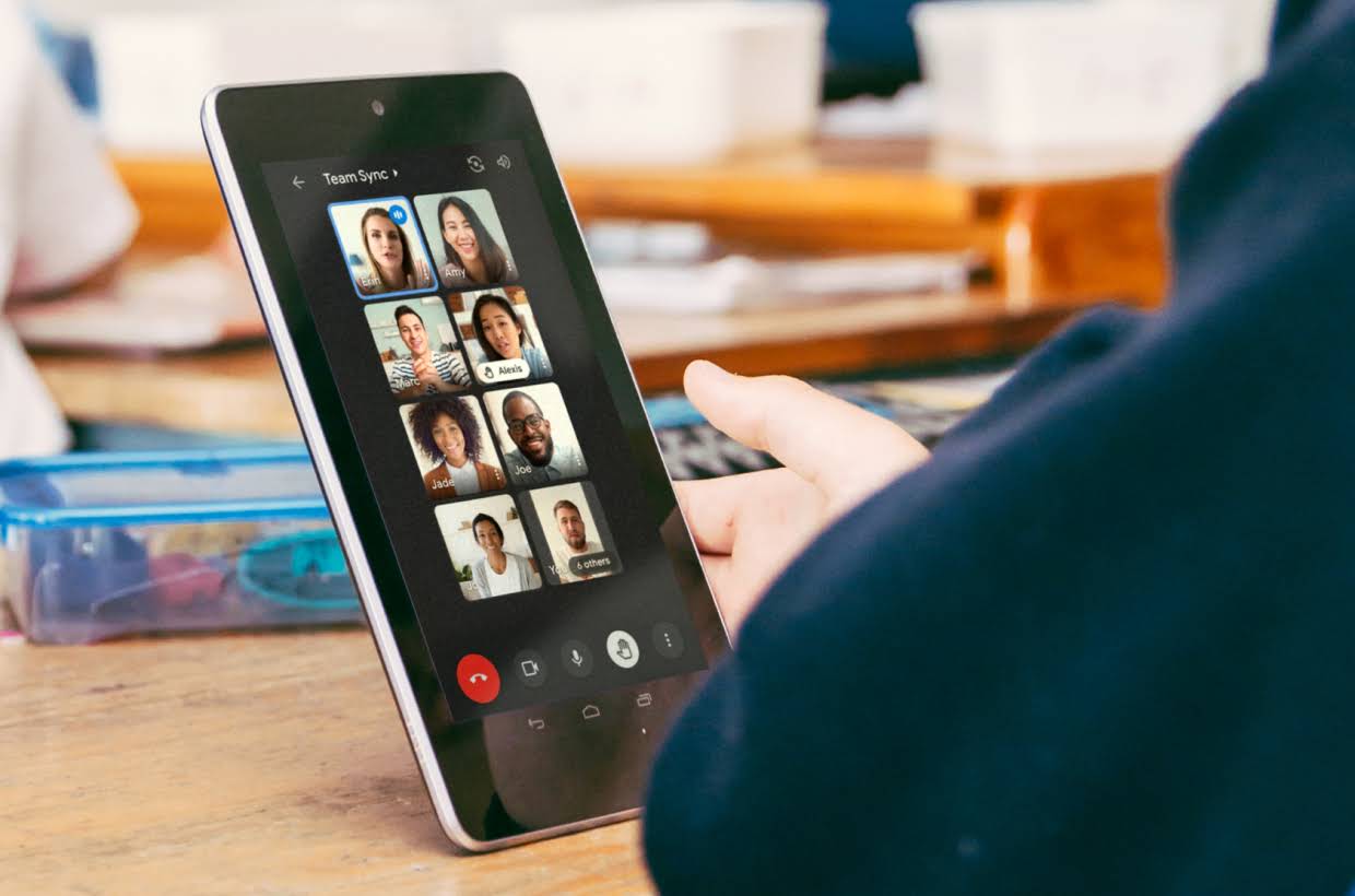 A hand on a table holding a tablet displaying a Google Meet call.