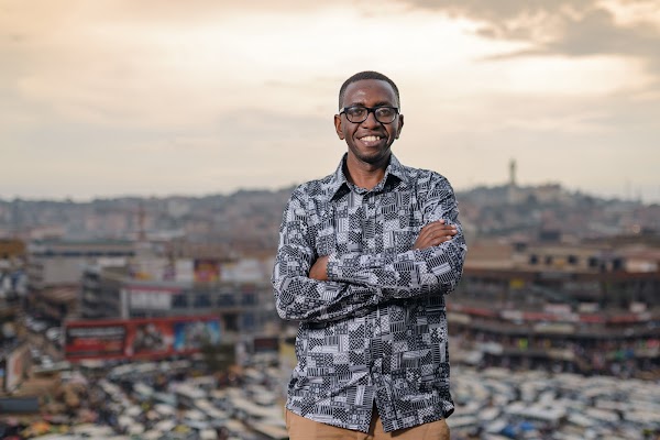 Un hombre con gafas y camisa de manga larga de camuflaje cruza los brazos sonriendo con una ciudad de fondo