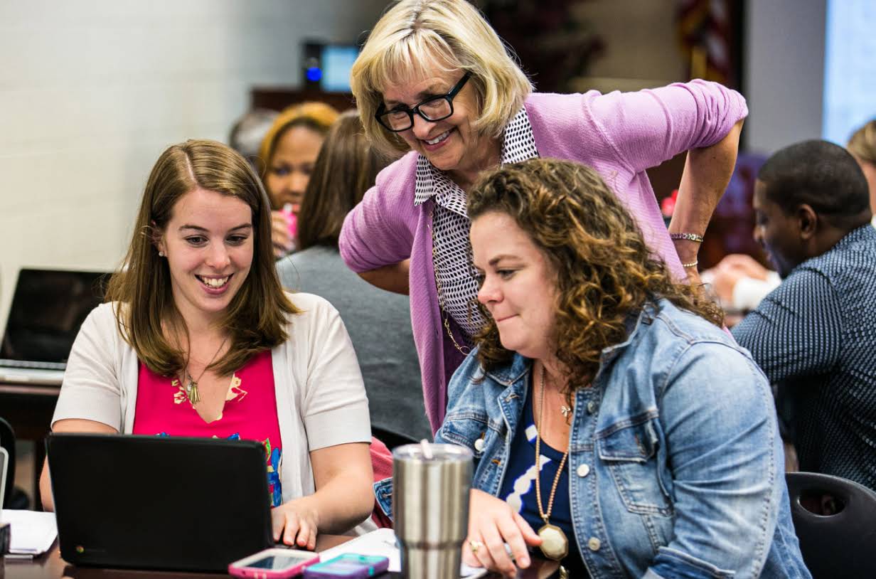 Trzy nauczycielki uśmiechają się, pracując na laptopie. Znajdują się w sali konferencyjnej wypełnionej ludźmi, którzy siedzą przy stołach i pracują na laptopach.