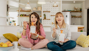 Two women sitting legs crossed on a couch, eating pasta out of a takeout container.