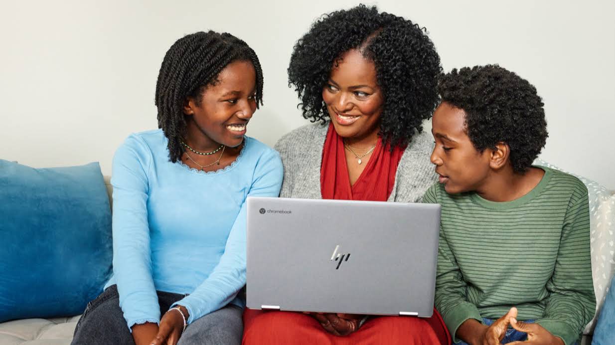 Una madre escribe en una Chromebook mientras está sentada en un sofá junto a dos hijos.