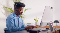 Una persona que usa audífonos con micrófono y una camisa con cuello azul está sentada en un escritorio y escribiendo en una computadora.