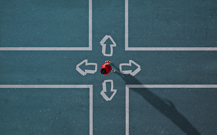 Decorative: woman dressed in red standing in the center of four arrows painted on a green asphalt pointing in four different directions, i.e., north, east, south and west.