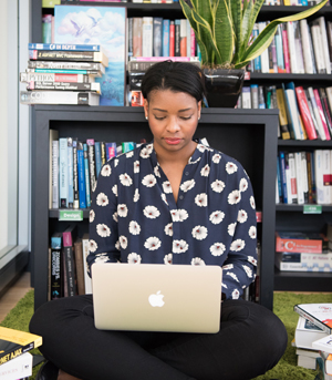 a woman sits on the floor in front of a bookcase on her laptop