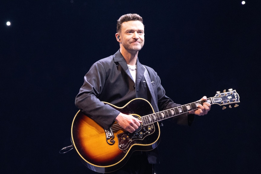 A man stands smiling on stage, strumming a guitar around his neck. He wears a grey button up shirt.
