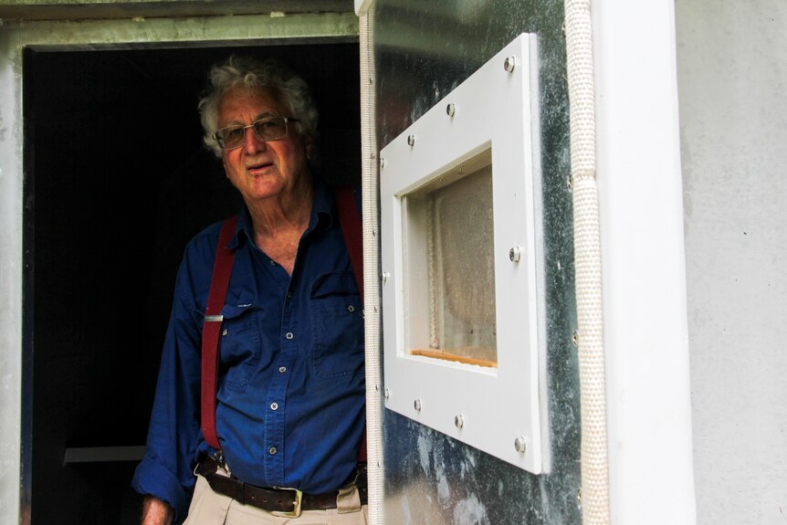 A man wearing a long-sleeved blue shirt and red braces stands, looking out of a bunker.