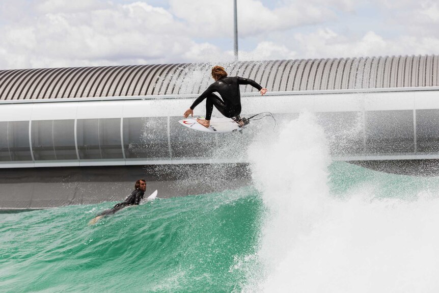 Two surfers, one lying on his surfboard and other wave jumping on top of a blue-green wave with a modern building behind them.