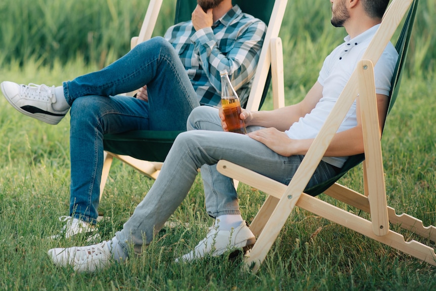 Two men chatting and holding beers while sitting on deck chairs in the grass in front of a field.