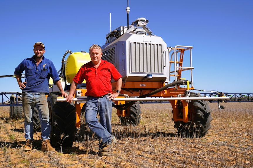 An older and young man stand next to tractor-looking robot in a paddock with stubble, blue skies.