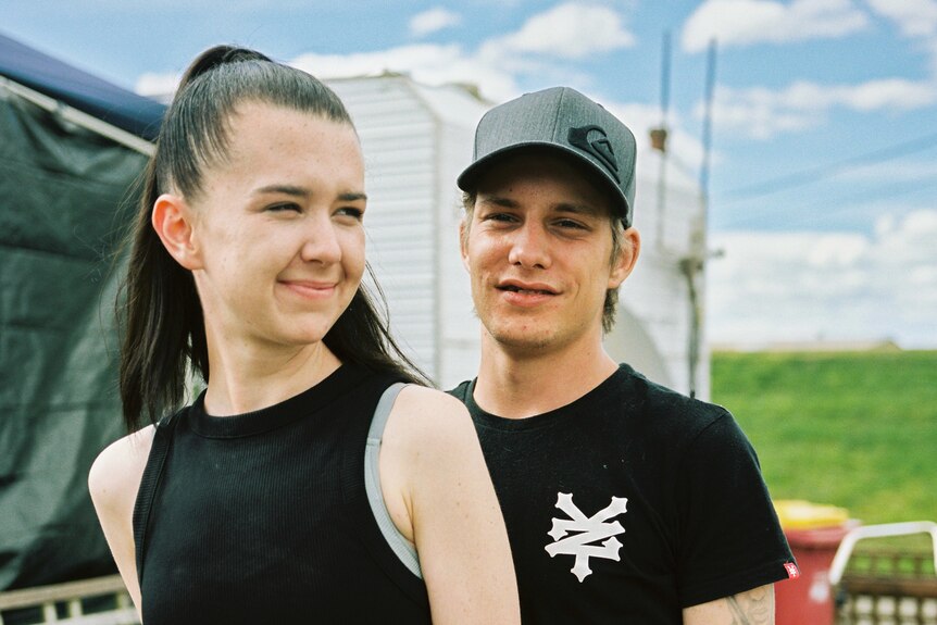 A young couple pose, the man has his arm around the woman's waist