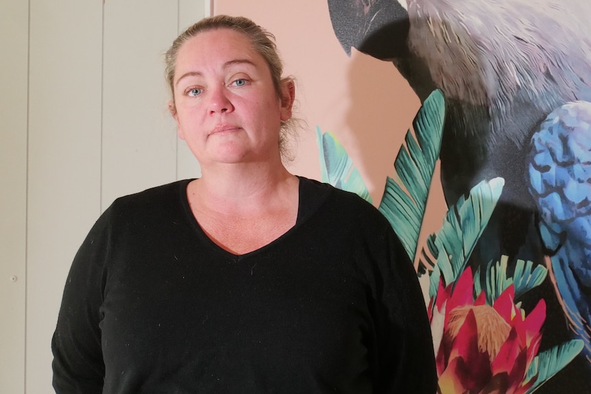 Woman standing staring at camera, with large artwork on wall behind her. 