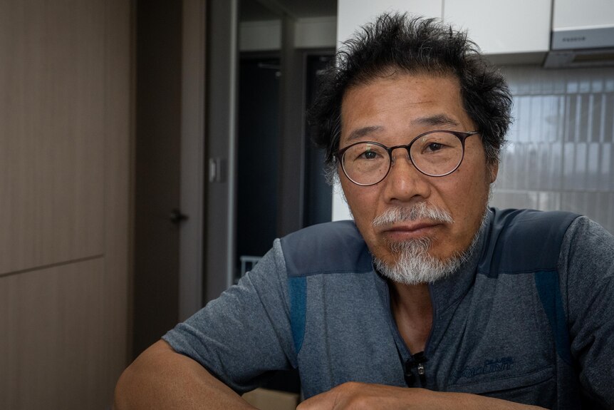 A bespectacled man with a grey goatee wears a blue top as he sits with his arms on a table in front of him.