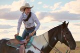 Tyler Morton lifting herself onto a black and white horse wearing jeans, white long sleeve top and a hat.