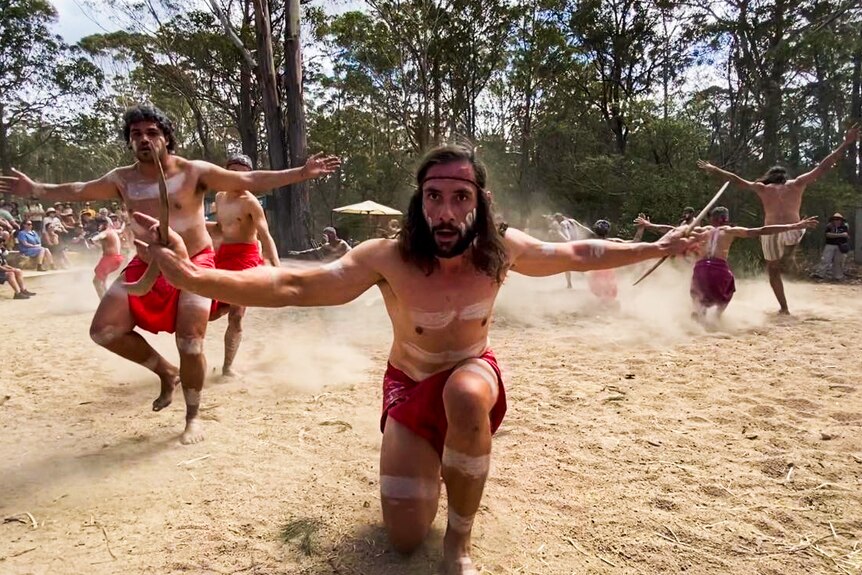 An Indigenous man wearing traditional clothes dances at the front of a circle of people gathered outdoors.