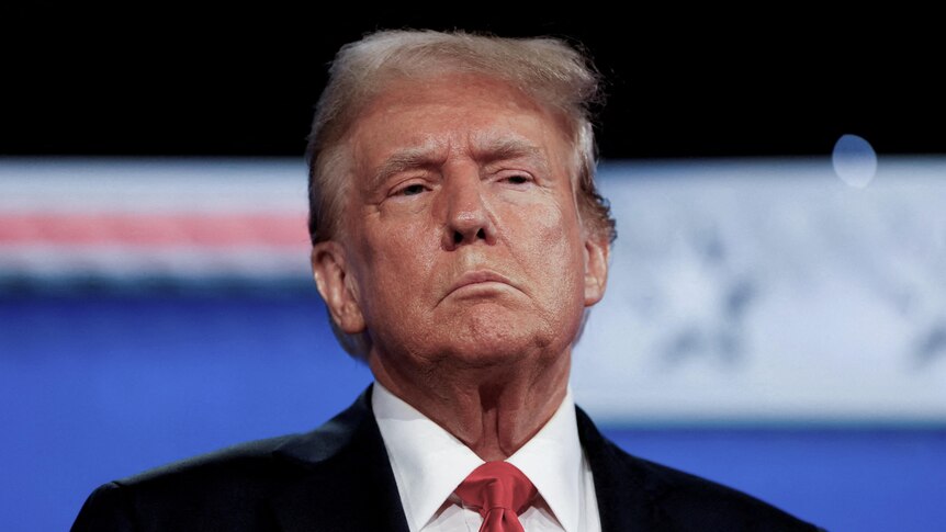 An older man wearing a red tie, white shirt and a black suit jacket with a serious expression