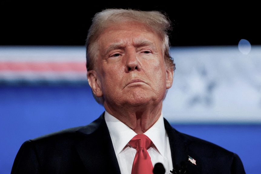 An older man wearing a red tie, white shirt and a black suit jacket with a serious expression