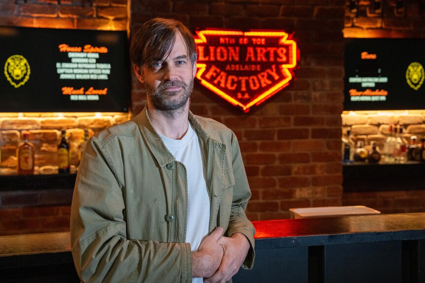 A man leans an arm against a bar top, behind him bottles of alcohol and a neon bar sign