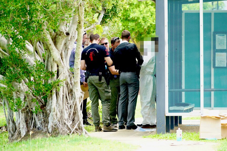 A group of detectives surround a man wearing a white forensic suit near a bus stop.