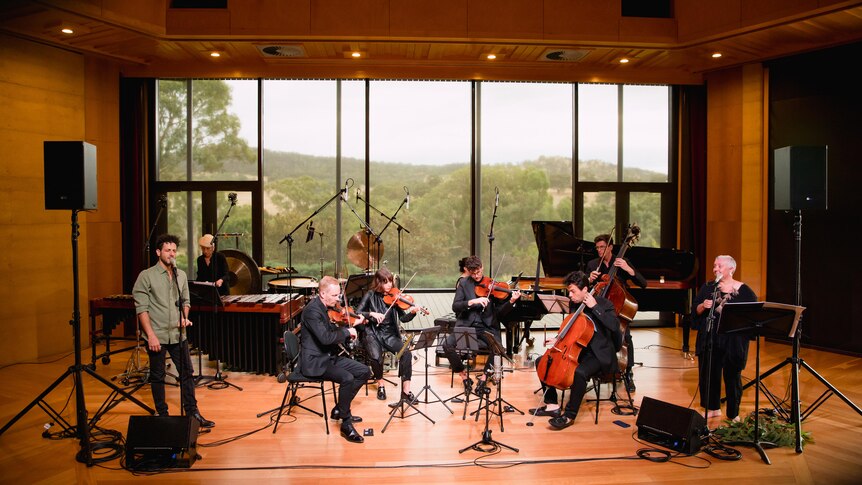 Lior and Dr Bennett on stage with musicians a large window in the background show Adelaide Hills bush.