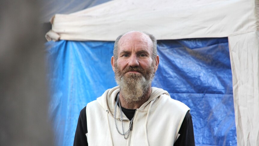 A bearded, balding man, stands in front of a makeshift home made of tarps and agzebos. 