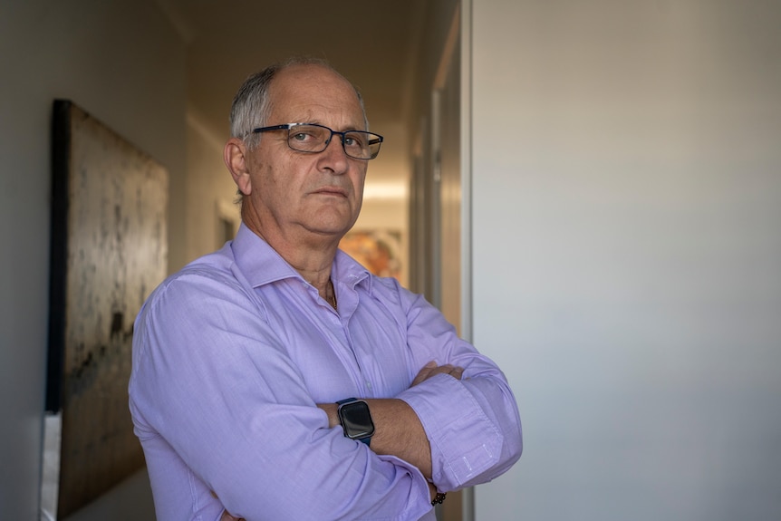 A man stands inside with his arms crossed, looking serious.