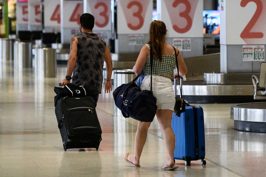 A young man and woman walk away with suitcases in from of empty baggage arrivals area.