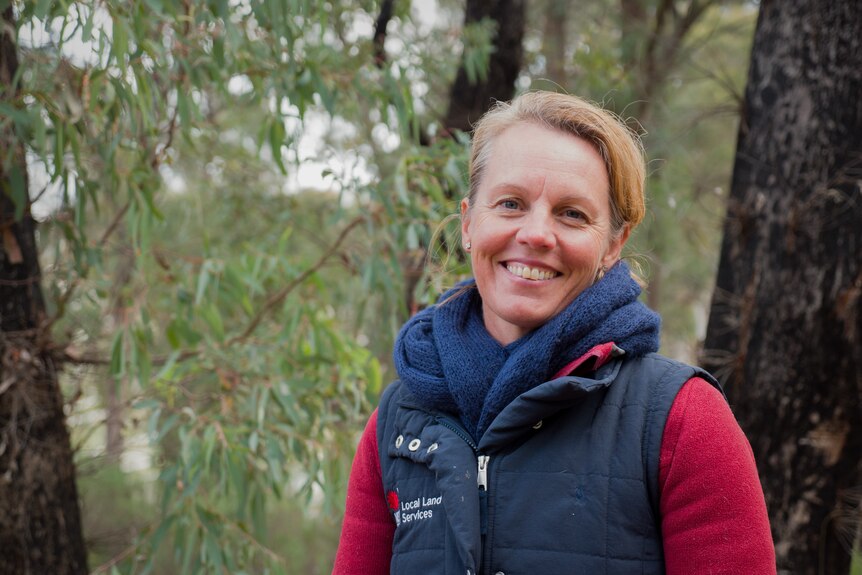 A woman with blonde hair in a bun smiles at the camera while wearing a navy vest atop a red sweater and standing in bushland.