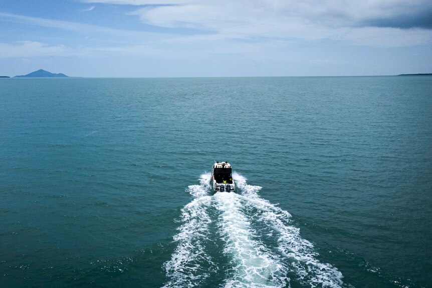 An aerial view from behind a speed boat shows it heading out on a clear day towards two land masses on the horizon