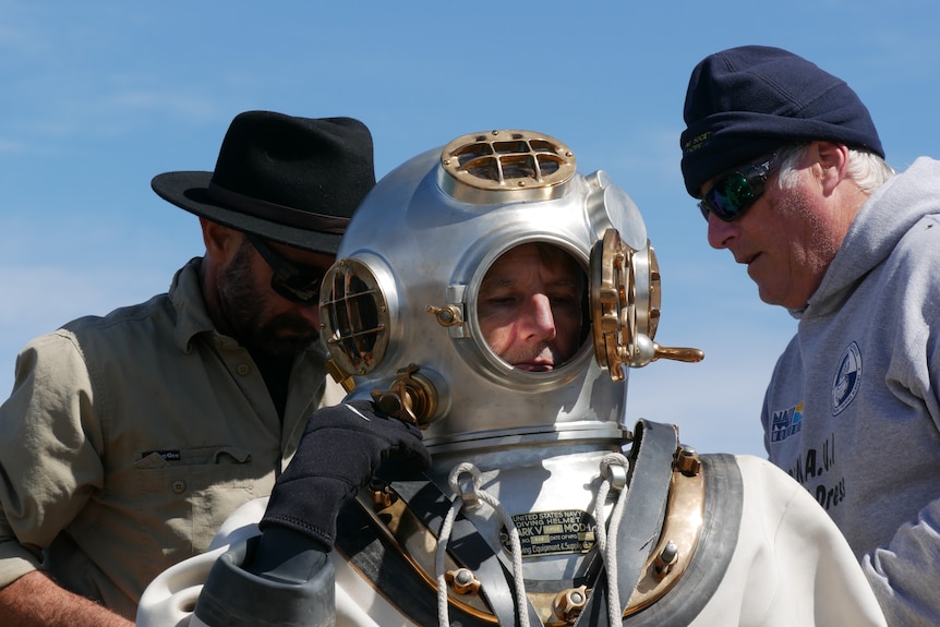 A close-up of a copper diving helmet with the window open as two men help tighten screws on the chest plate.