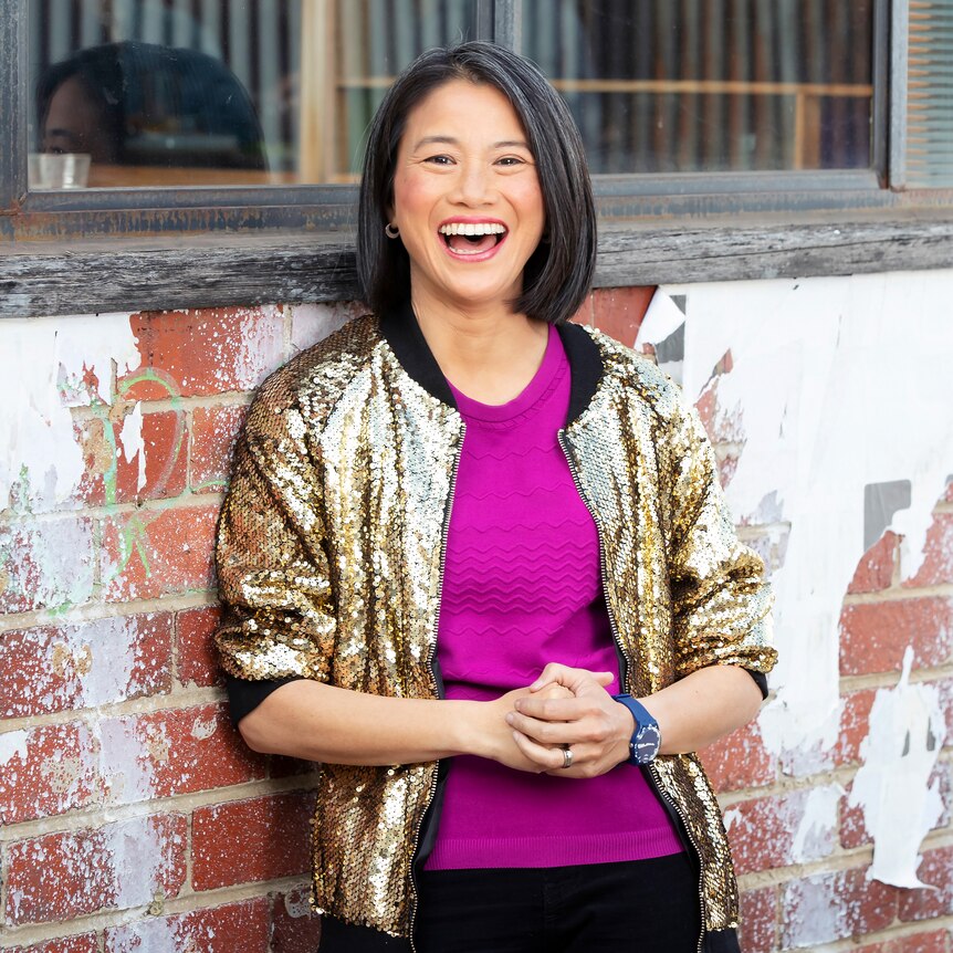 Lisa Leong leaning against a brick wall with windows in it, laughing.