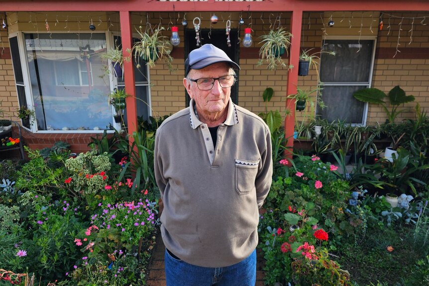 A mean wearing a grey sweater, glasses, and baseball cap standing in front of a brick house with garden