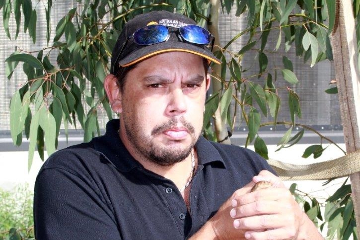 An Aboriginal ma wearing a hat and black shirt stands with his hands clasped among Eucalyptus leaves and branches. 