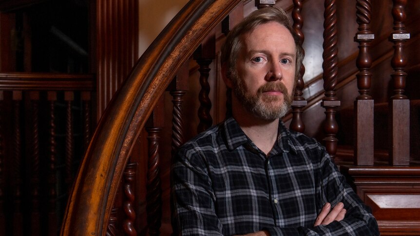 A white, middle-aged man with a sandy hair and beard stands with his arms crossed in a timber-panelled room