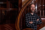 A white, middle-aged man with a sandy hair and beard stands with his arms crossed in a timber-panelled room