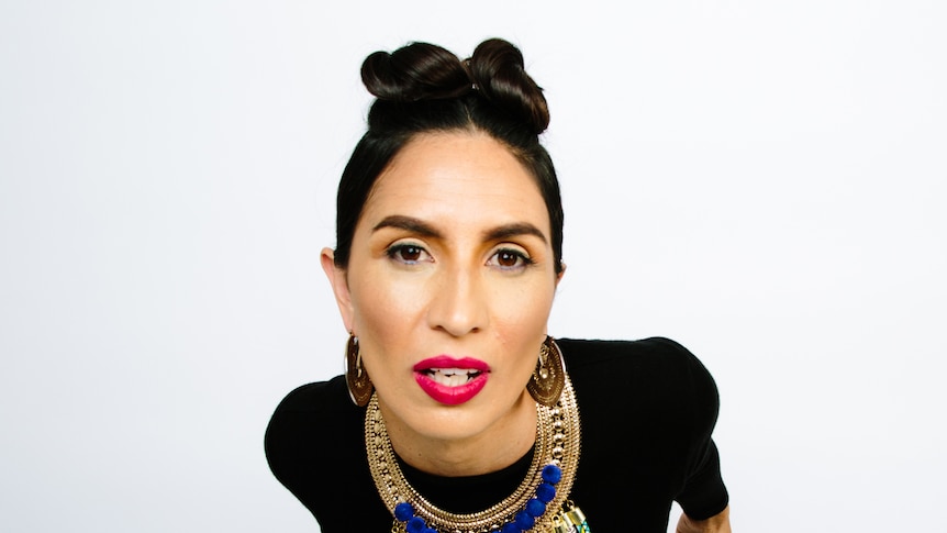 Maya Jupiter wearing a large beaded necklace standing in front of a white background leaning in towards the camera