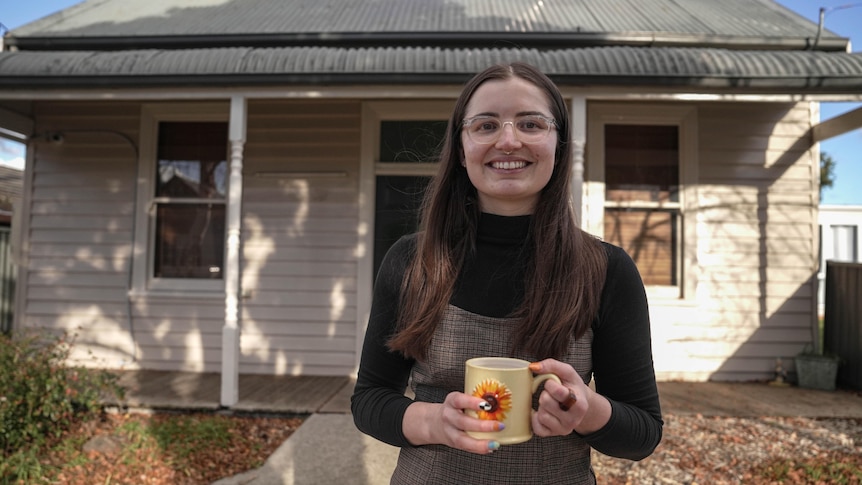 A young woman wearing grey dress over black skivvy stands in front of a house, smiling with a mug in her hands