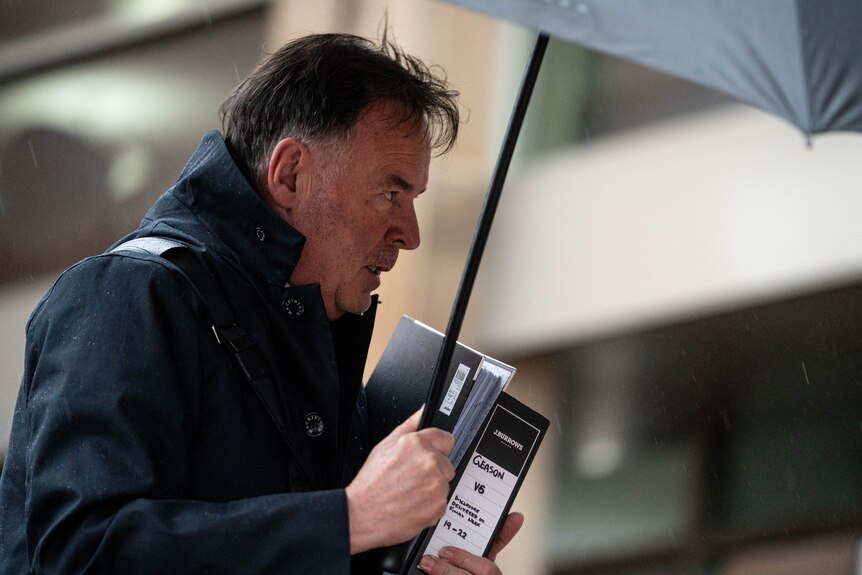 Man walking into court holding documents and covering his face with an umbrella.