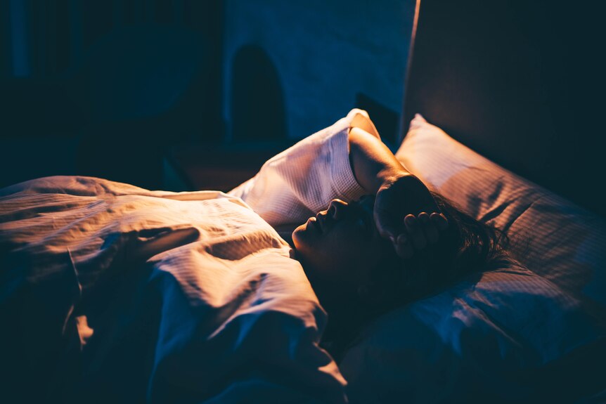 A young woman lies in bed with her hand on her forehead, unable to sleep