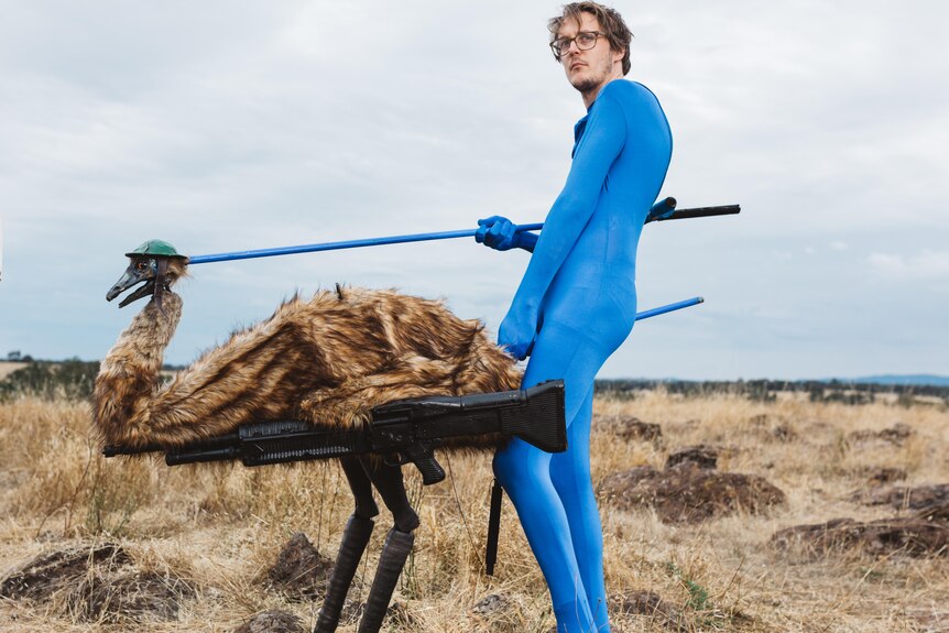 A man in a blue morph suit controls an emu puppet in a large bush field