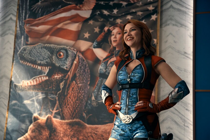 A woman in a superhero costume stands in front of a painting of herself riding a dinosaur